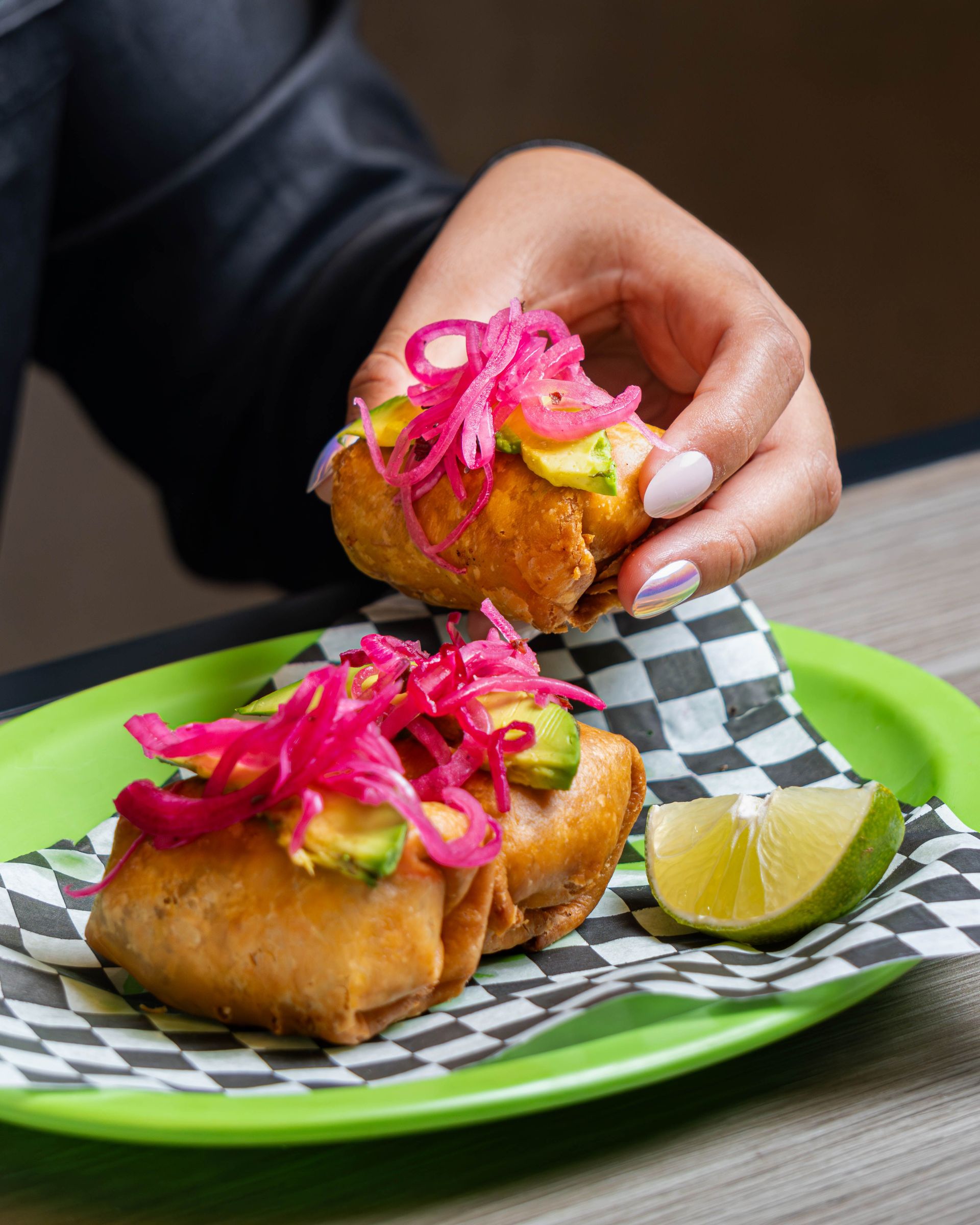 A hand holding fried tacos topped with pickled onions and avocado slices on a vibrant plate.